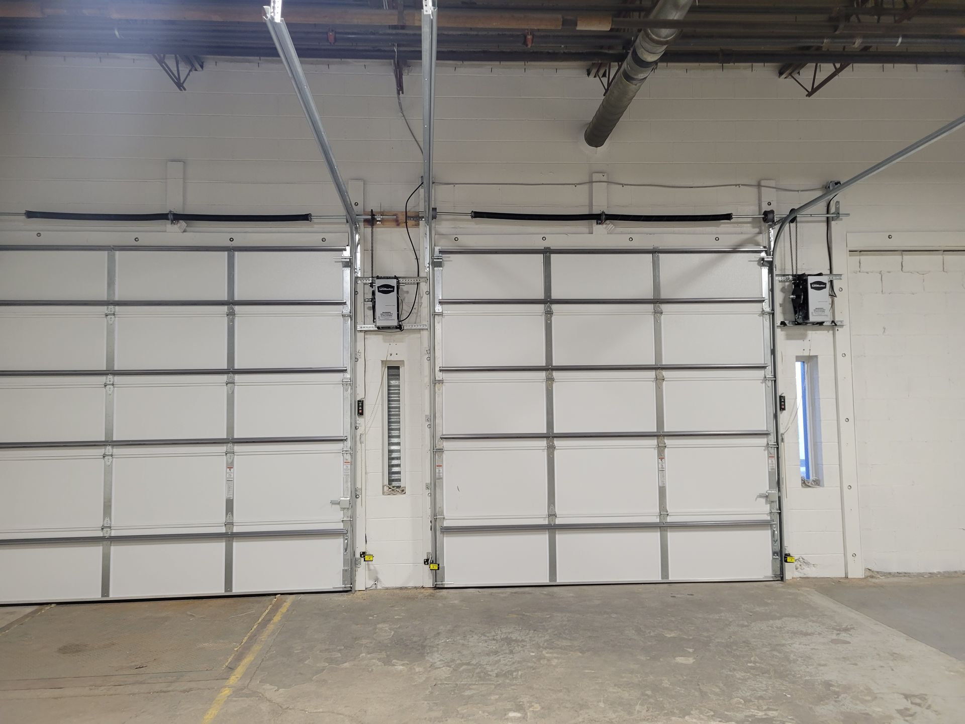 Two large white industrial garage doors side-by-side with wall-mounted controllers and narrow windows on a white wall.