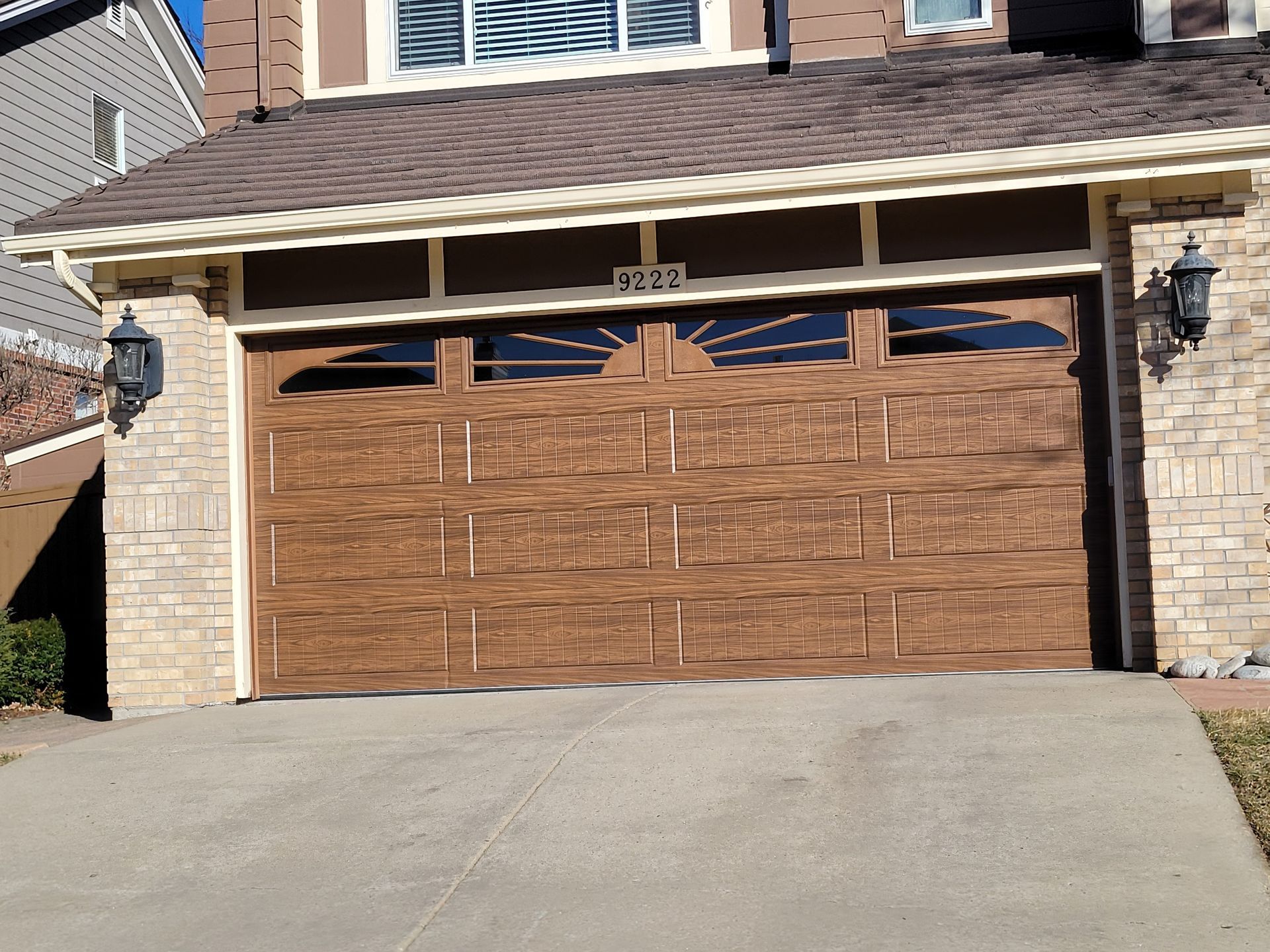 Brown garage door with arched decorative windows, set in a stone facade with exterior wall sconces.