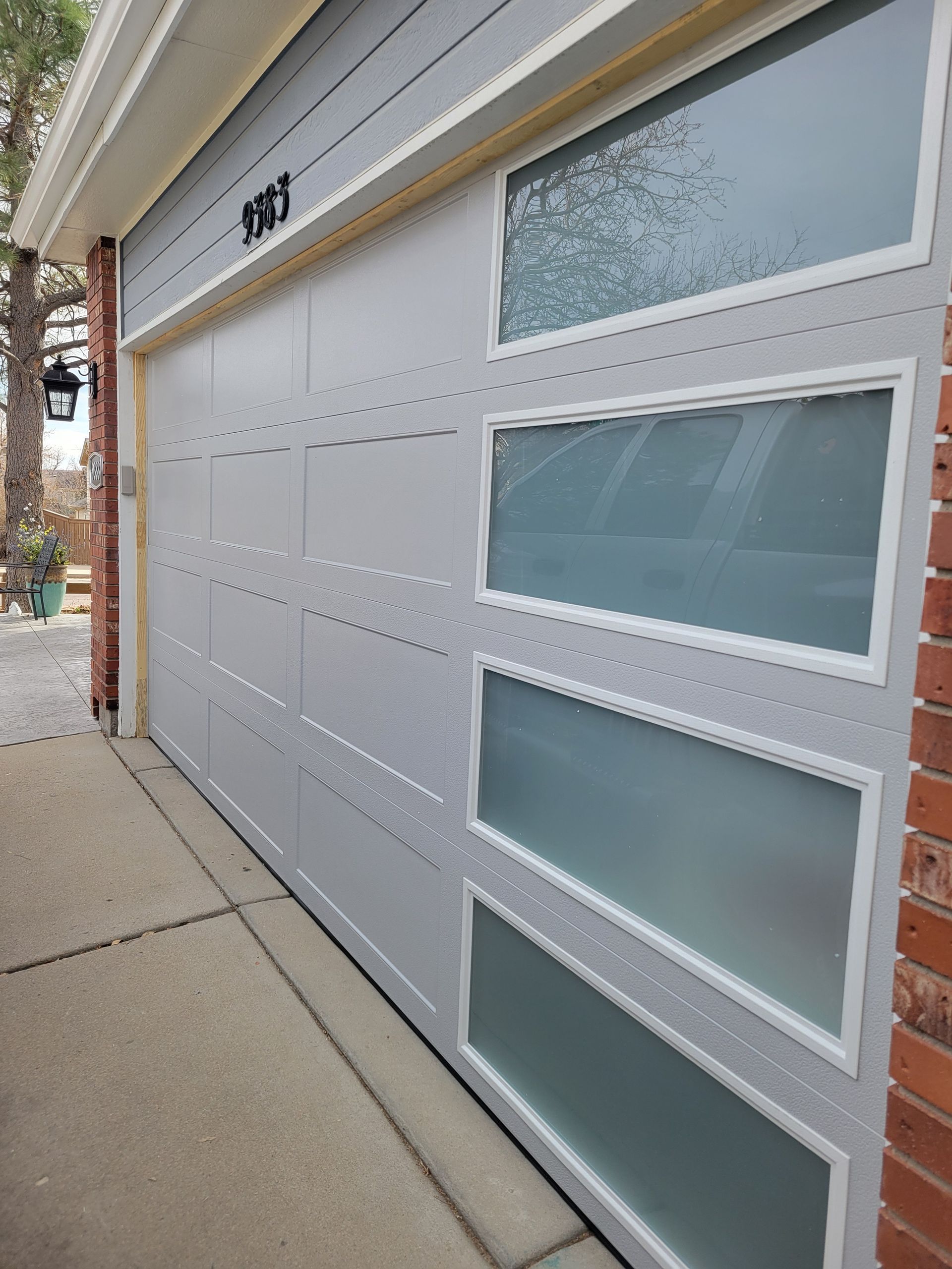A light gray garage door with a vertical row of four frosted glass windows on a brick house exterior.
