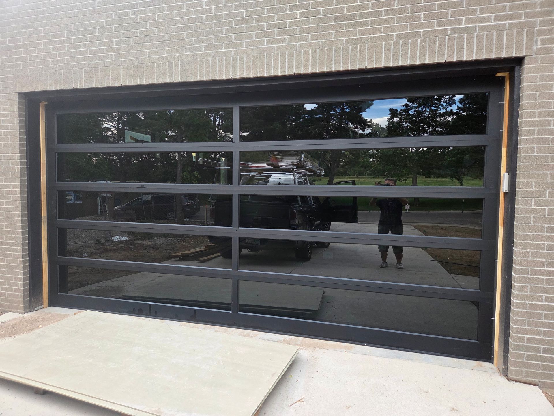 A modern, black-framed glass garage door installed in a light brick exterior wall, reflecting a vehicle and surroundings.