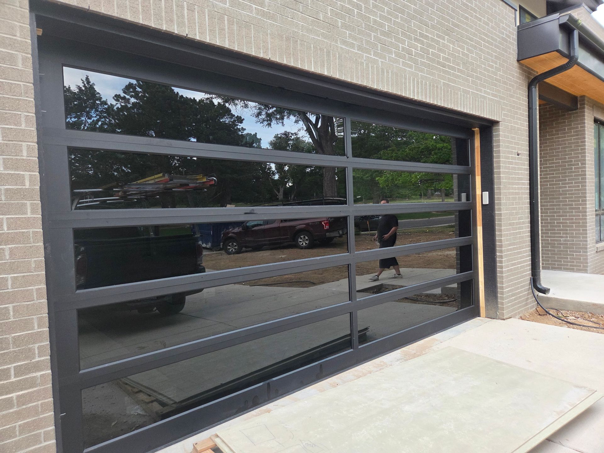 A modern, black-framed garage door with horizontal glass panels reflecting a driveway and trees against a brick house.