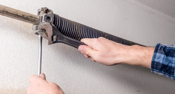 A person uses an adjustable wrench and a metal winding bar to tighten a garage door torsion spring.