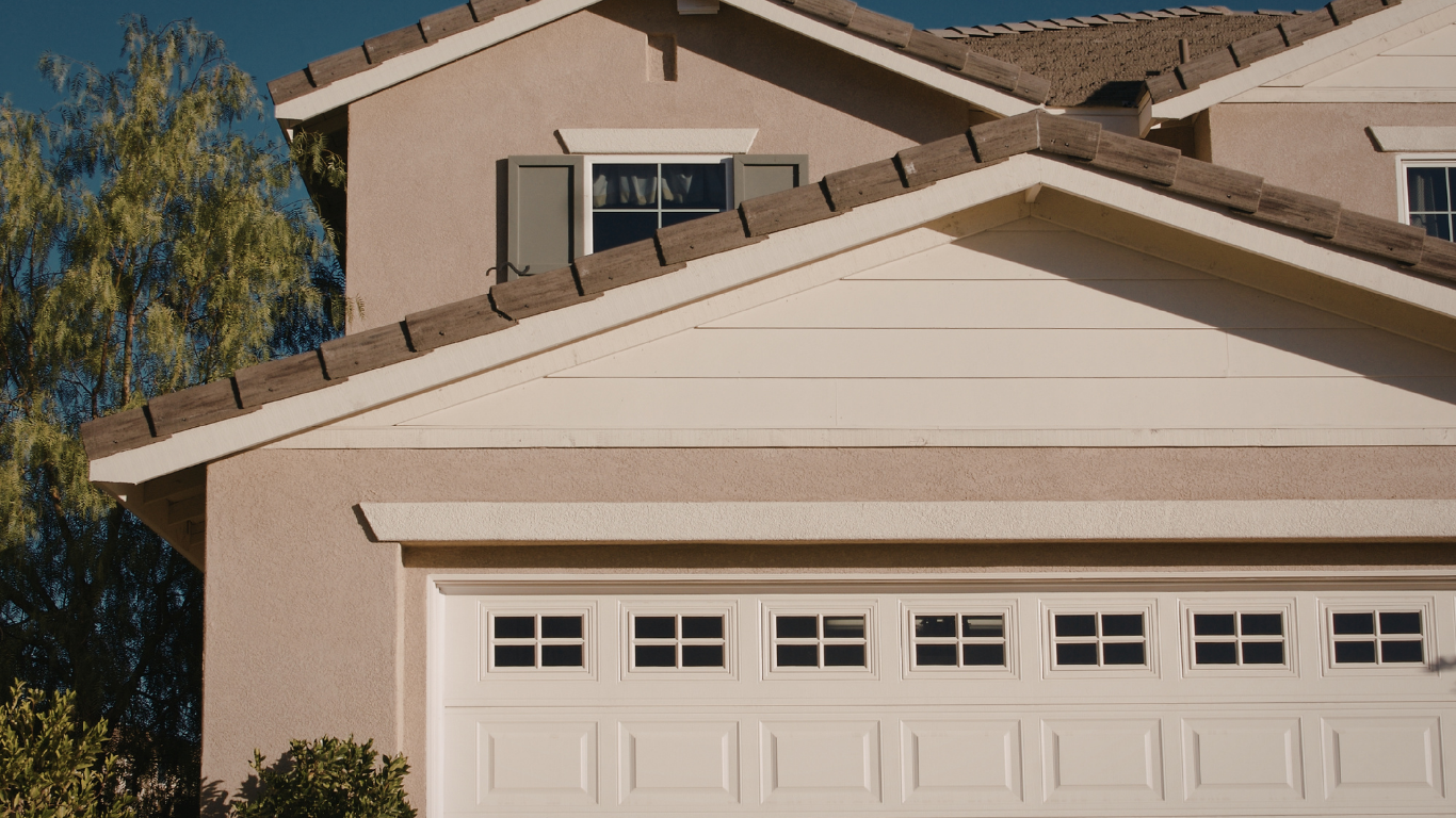 A beige two-story house with a white garage door, brown roof tiles, and a window with gray shutters.