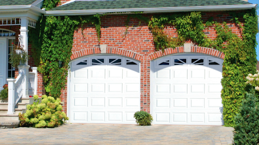 A brick home with two white, arched garage doors featuring decorative windows, surrounded by ivy and landscaping.