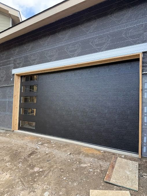 A modern black garage door with a vertical row of five rectangular windows installed on a house under construction.