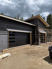 A modern home under construction with black siding, a horizontal-paneled garage door, and scaffolding in a wooded area.