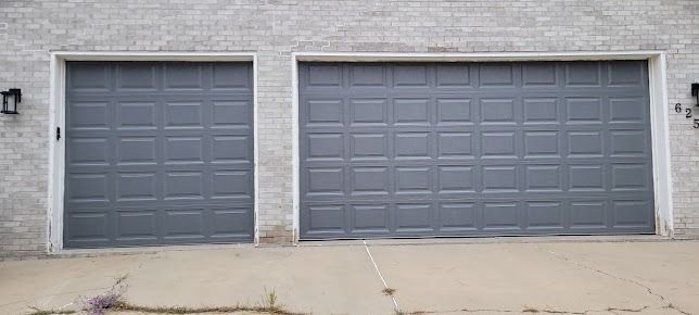 Two charcoal gray garage doors on a light brick house facade with a concrete driveway in the foreground.