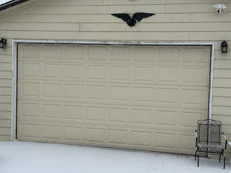 Beige garage door with a black eagle ornament above it, positioned next to a metal patio chair in light snow.