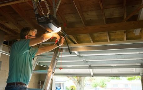 A person on a ladder repairs a garage door opener motor mounted to the ceiling.