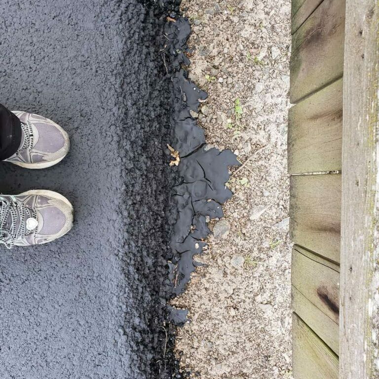A person 's feet are standing on a asphalt road next to a wooden fence
