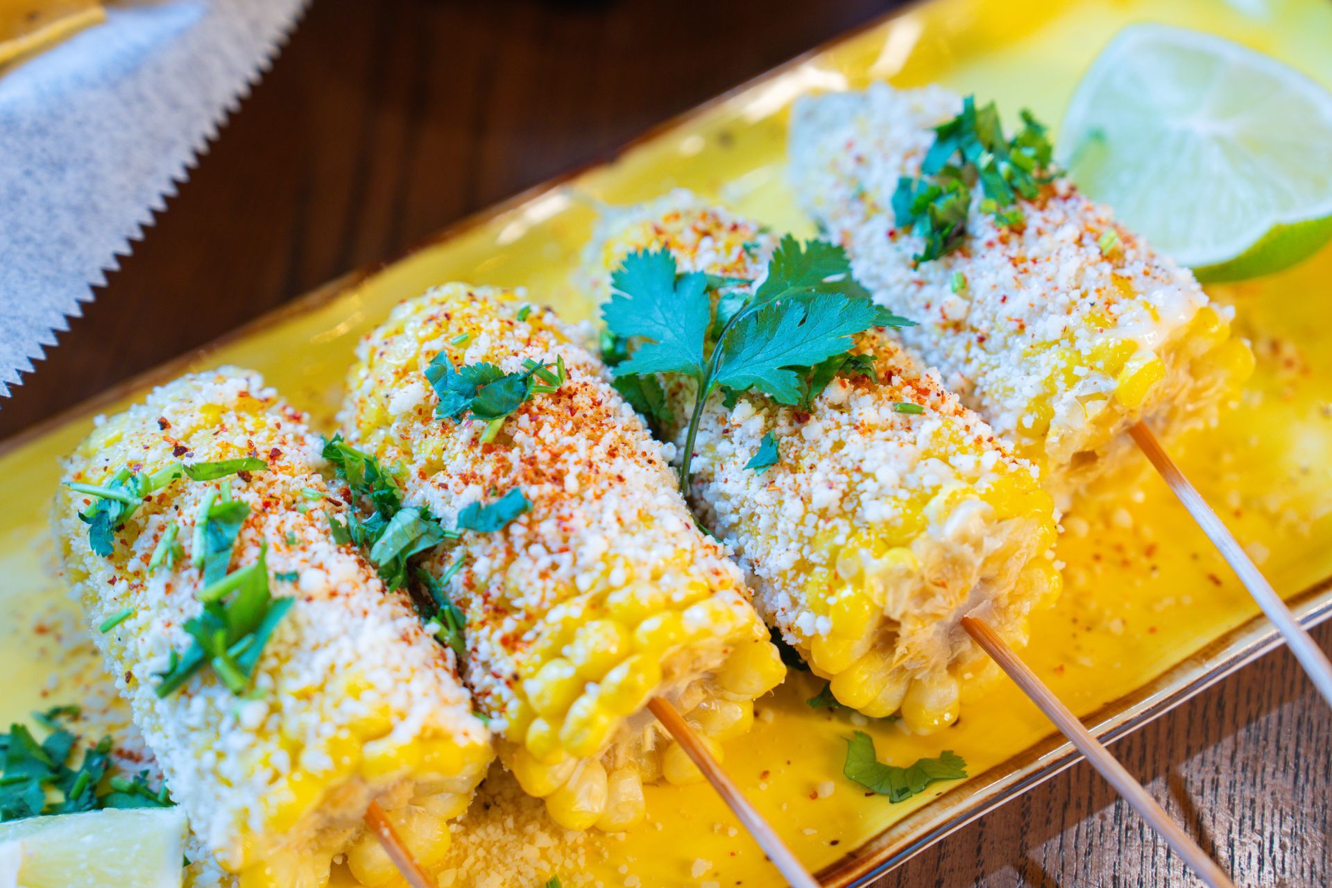 Four ears of grilled street corn on sticks, garnished with herbs and spices, sit on a red platter with lime wedges.