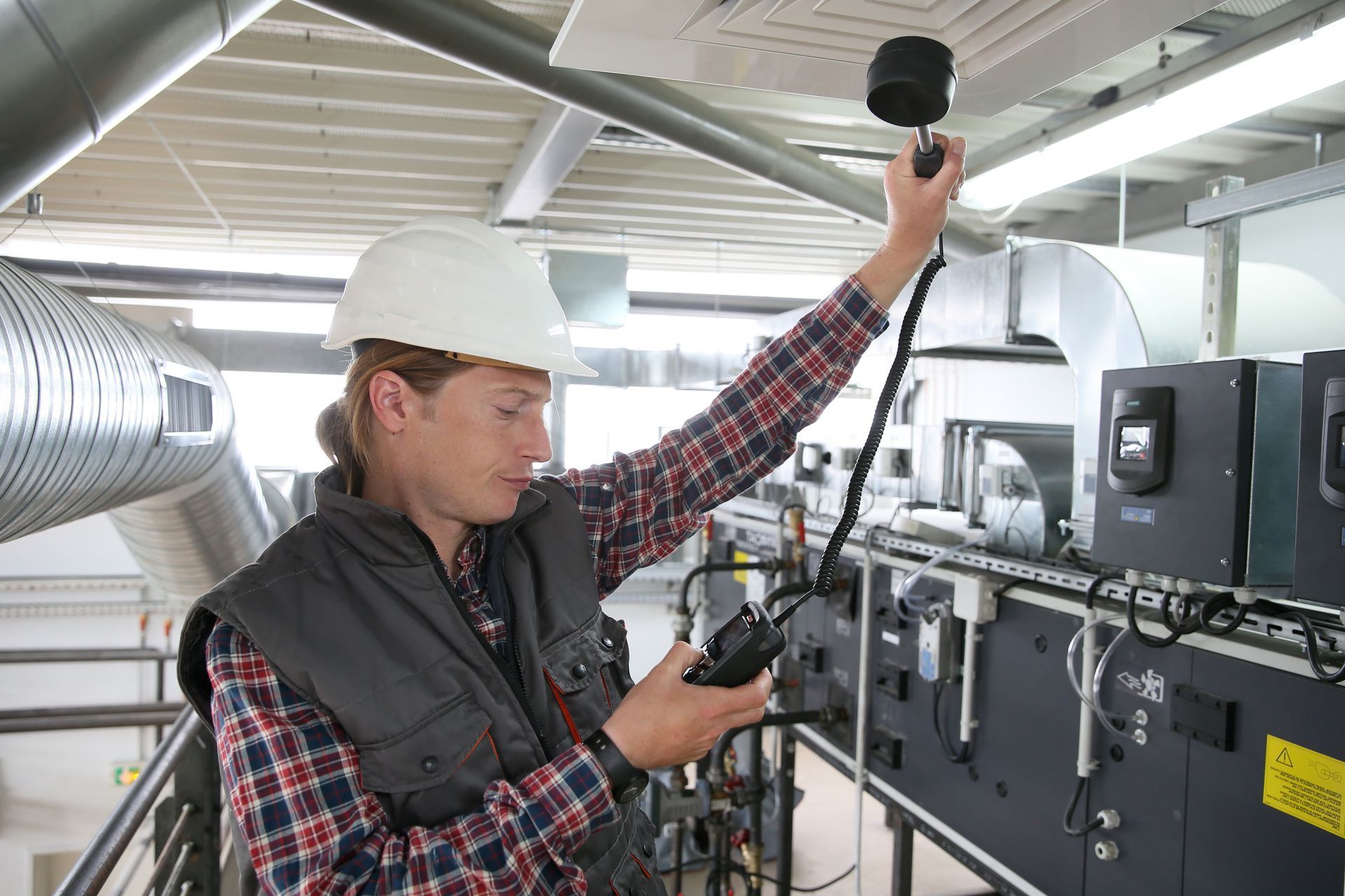 A man in a hard hat is holding a device in a factory.