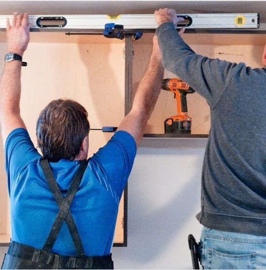 Professionals aligning and leveling cabinet shelves during installation. Two workers installing or measuring a cabinet shelf, one holding a level tool.