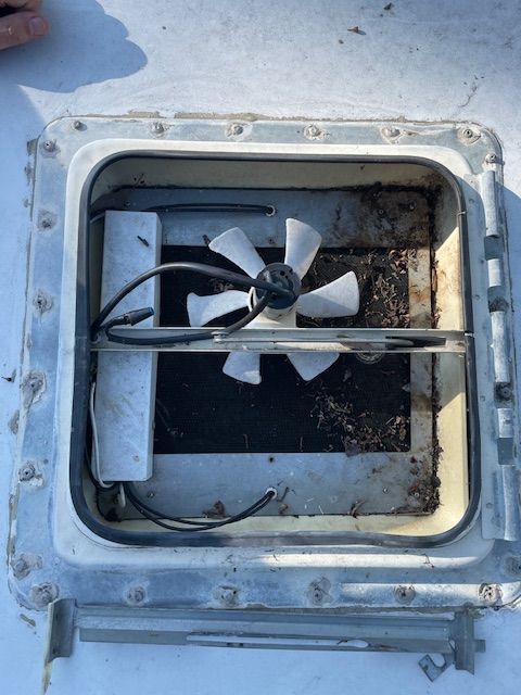 Open RV roof vent with fan, covered in debris. Gray metal, white fan blades, black wiring.