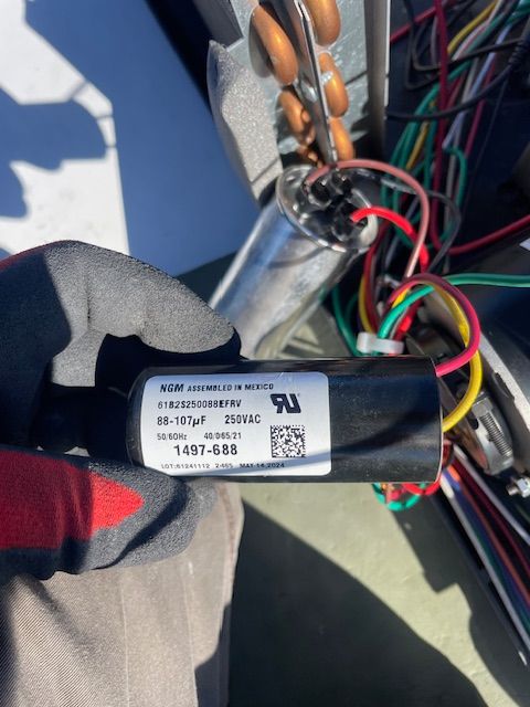 Gloved hand holds a capacitor next to air conditioner wiring; black and white label visible with specs.