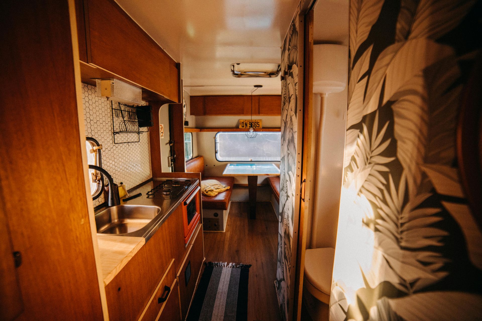Interior of a renovated camper van with wood paneling, kitchen, dining area, and leaf print curtains.