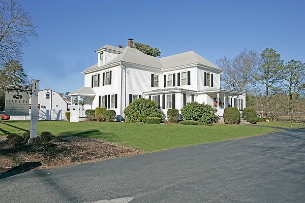 White colonial-style house with black shutters on a landscaped lawn beside a paved road