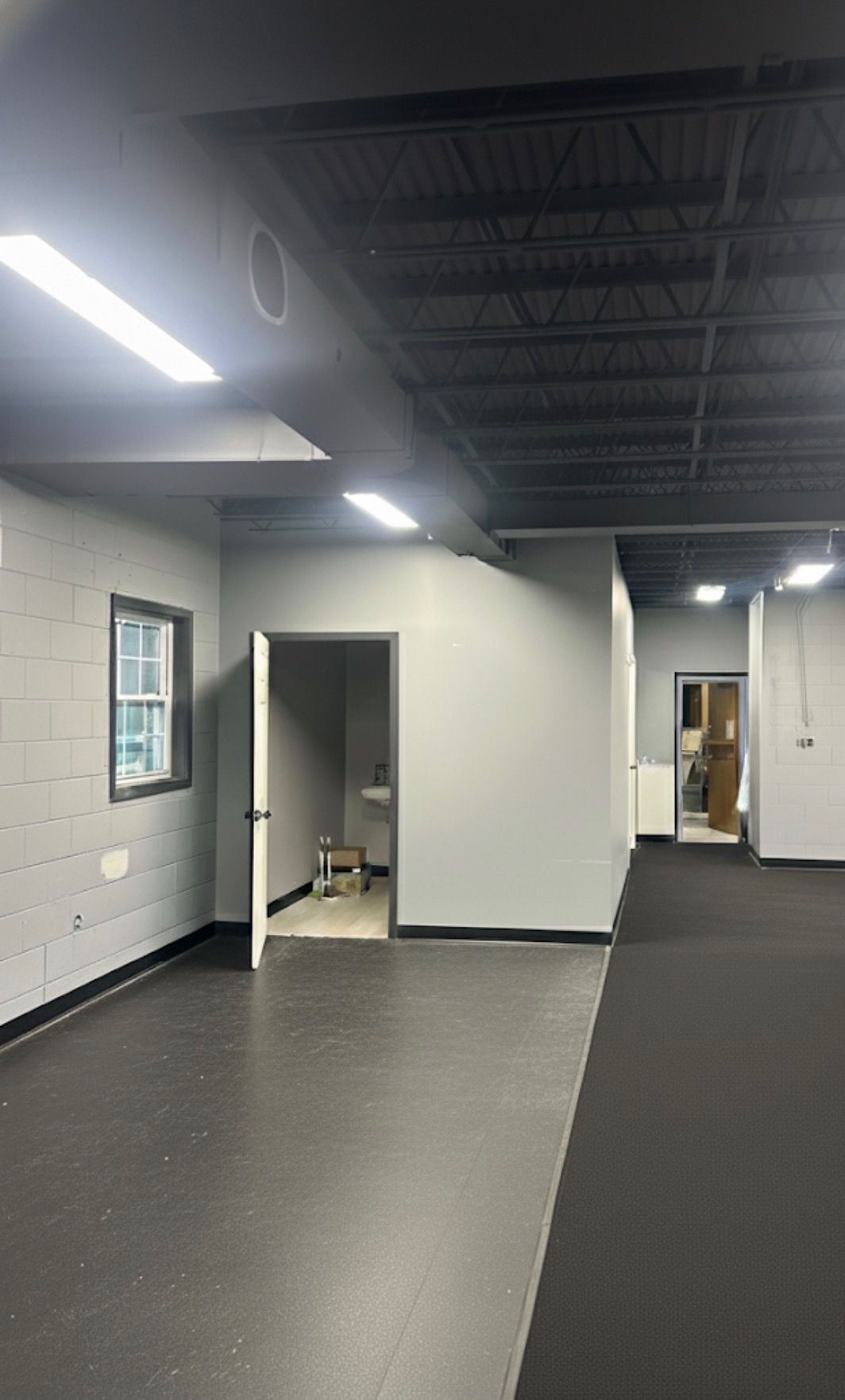Interior view of a gray-painted room with black flooring, a small bathroom, and fluorescent lighting.