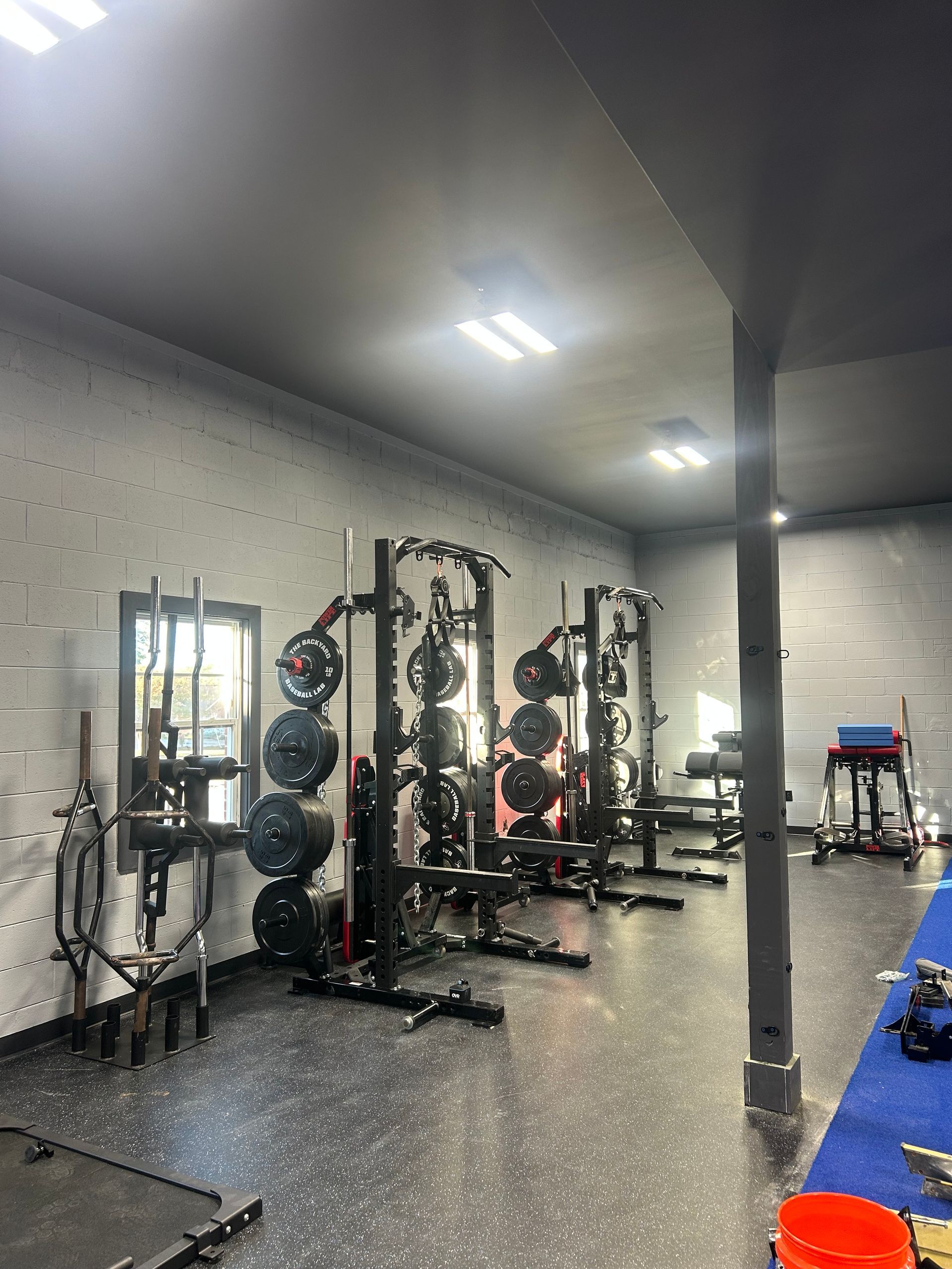 Gym with weightlifting racks, weights, and equipment on speckled flooring. Gray walls and ceiling.