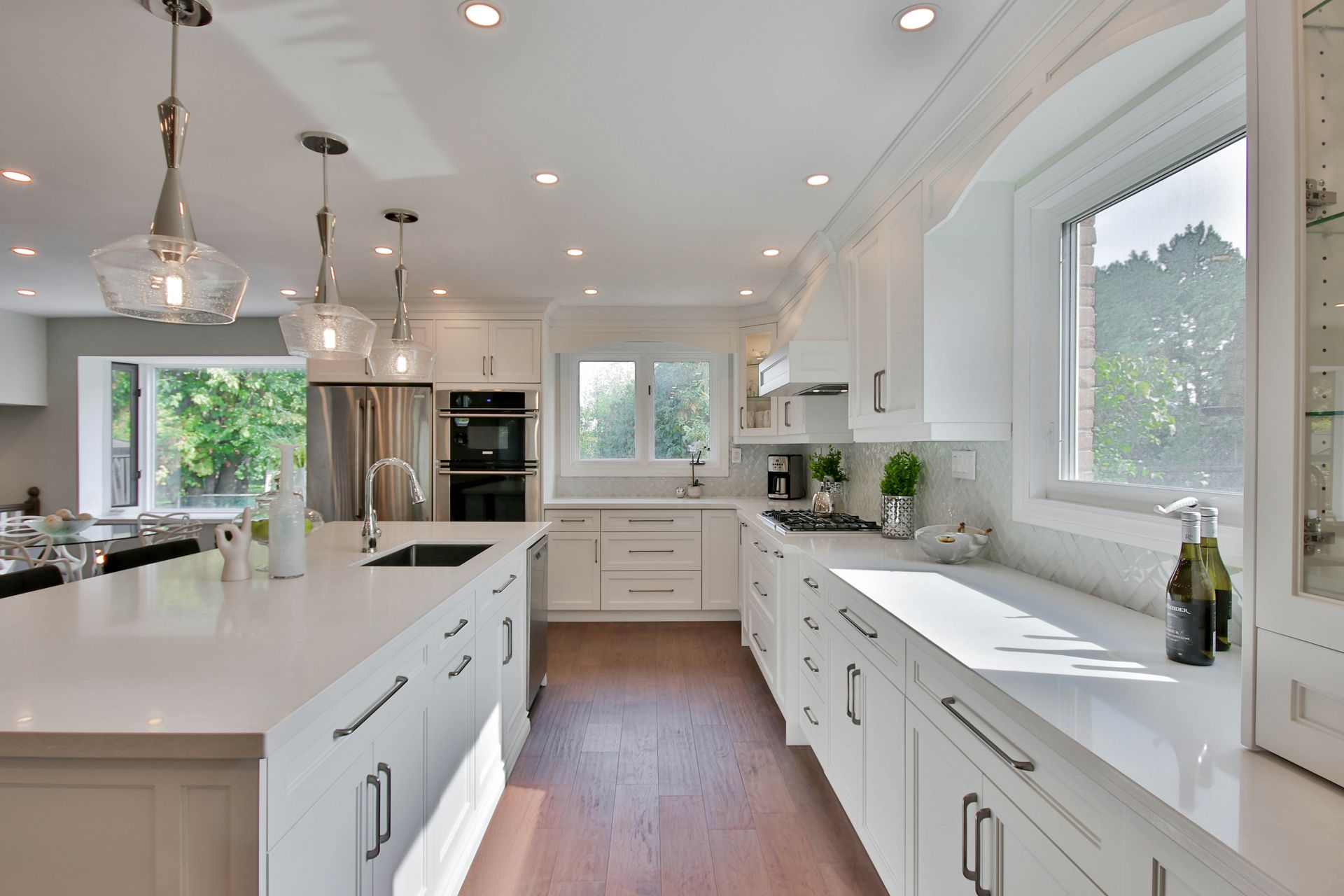 Bright white kitchen with island, cabinets, appliances, and windows overlooking greenery.