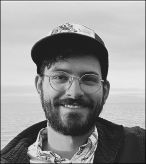 a black and white photo of Sam Schroeder smiling in front of the sea.