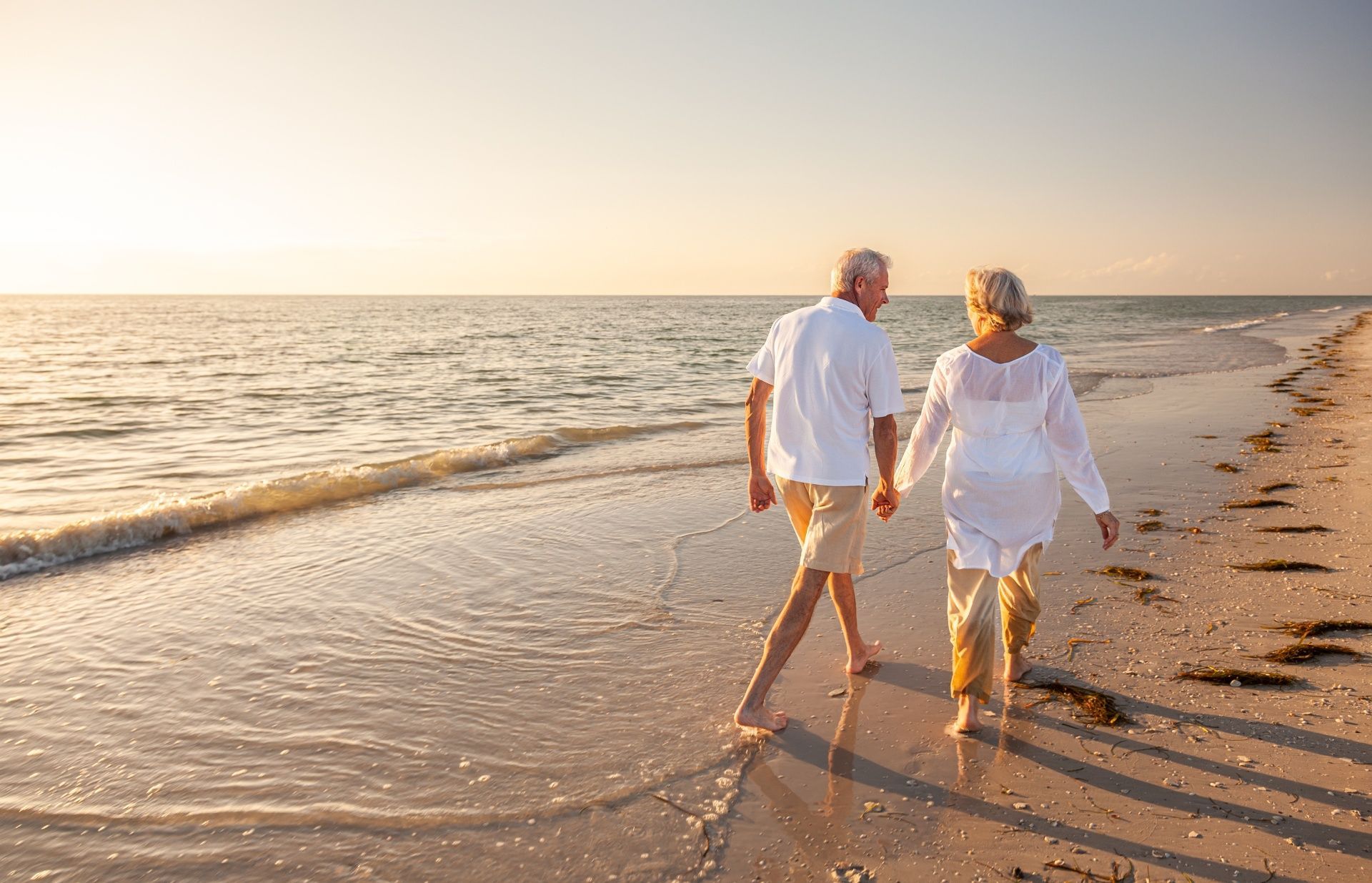 An elderly couple is walking on the beach holding hands.