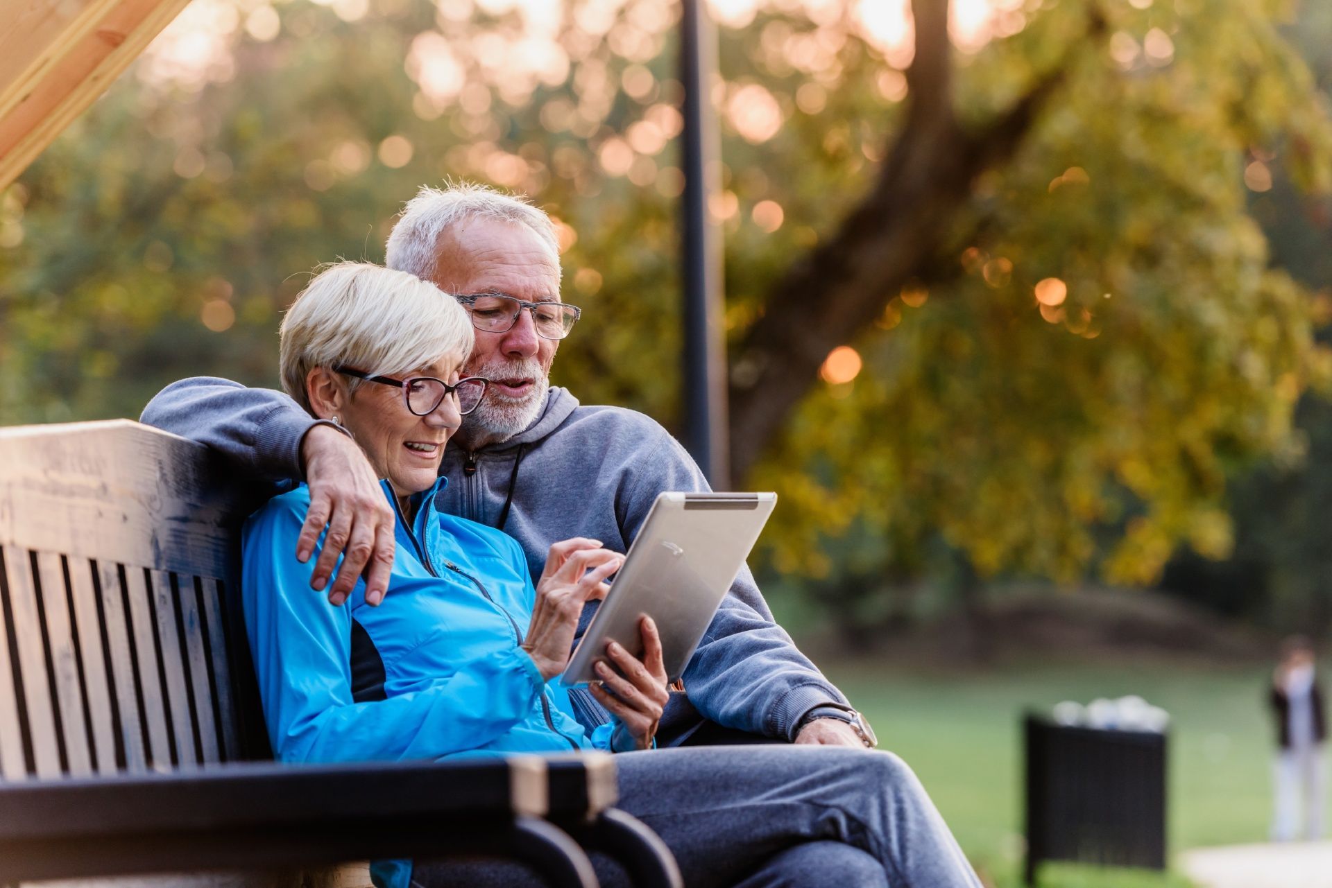 An elderly couple is sitting on a bench looking at a tablet.