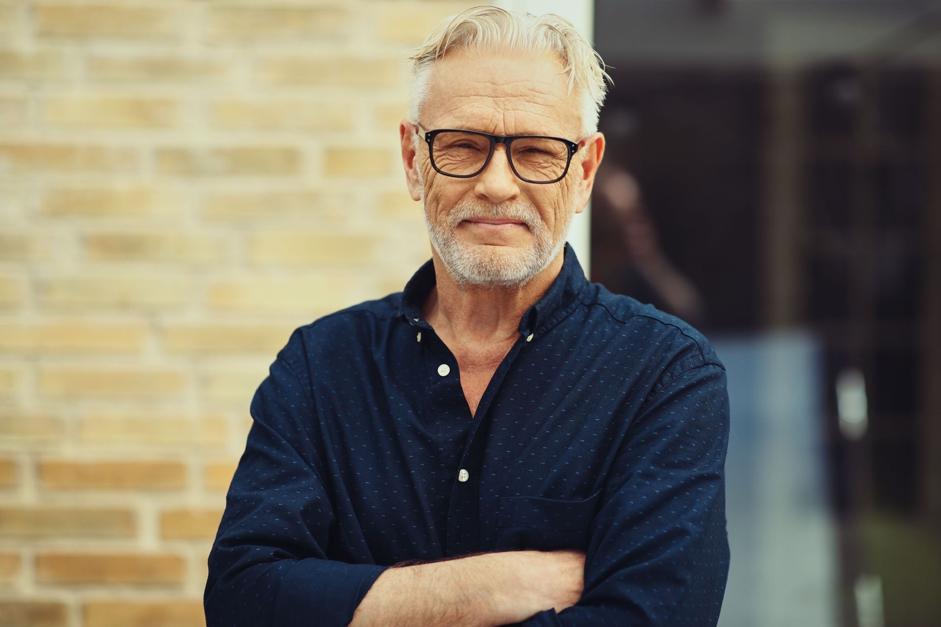 A man with glasses and a beard is standing with his arms crossed in front of a brick wall.