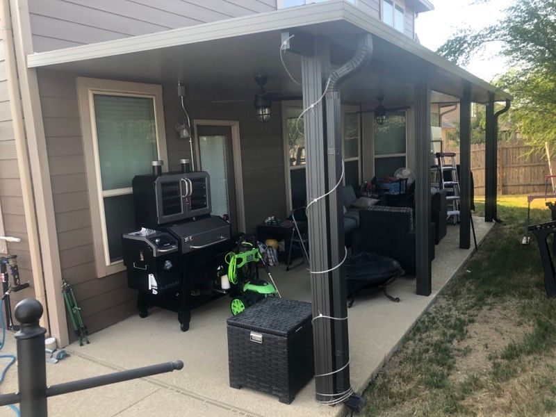 A covered patio with a grill , chairs , and a ladder in the backyard of a house.