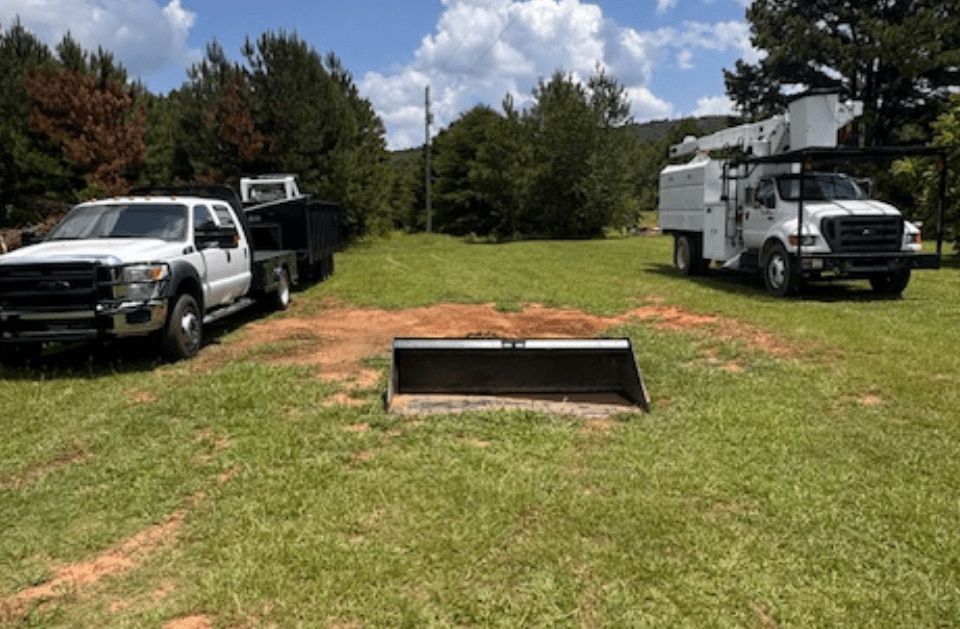 Three work trucks parked on grass, with a metal scoop in the foreground, trees and a blue sky in the background.