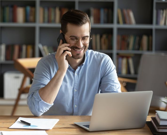 A man is sitting at a desk with a laptop and talking on a cell phone.