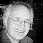 Lachende oudere man met bril, grijs haar, in een ruimte met zacht licht, mogelijk binnenshuis.