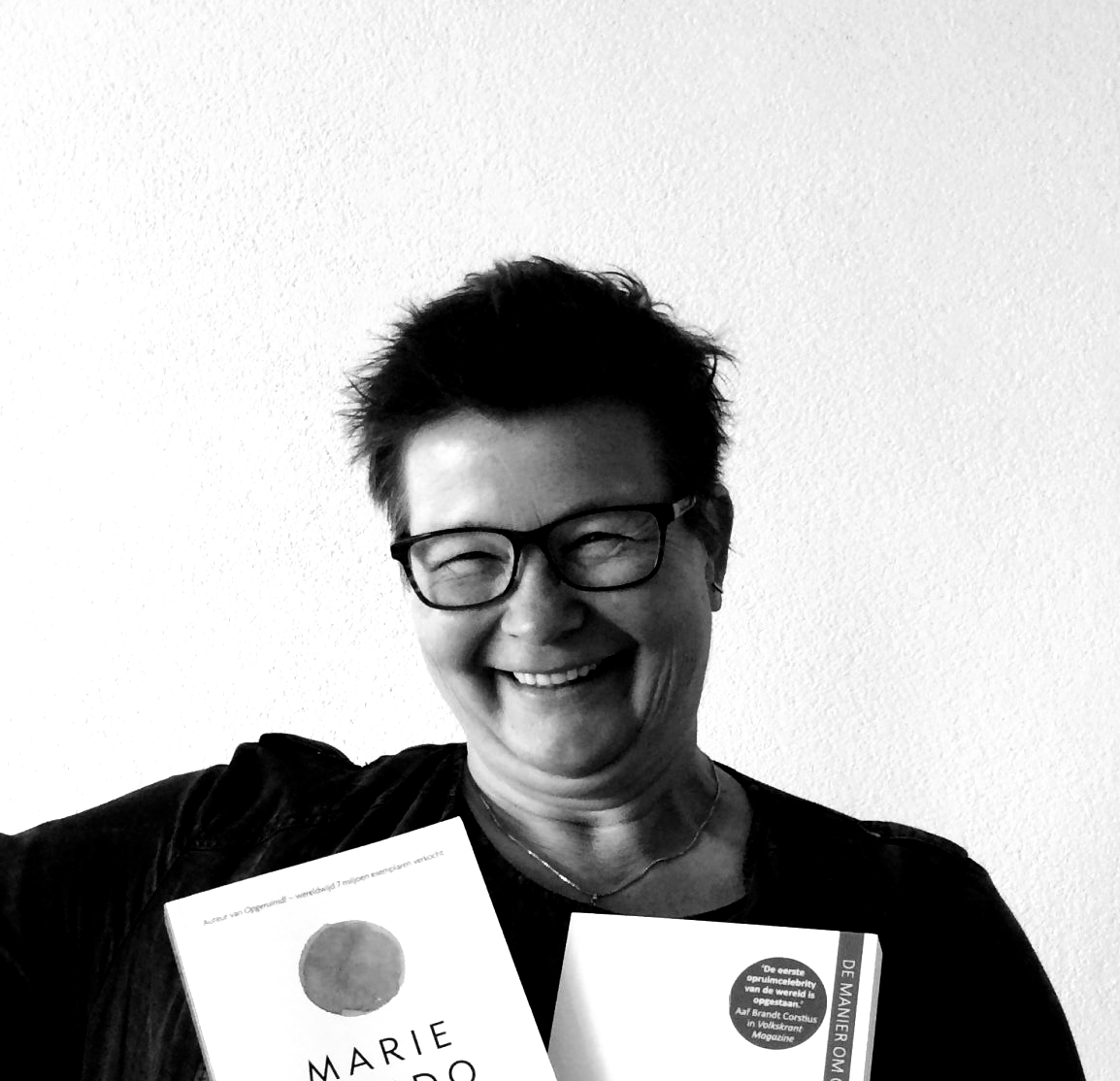 Lachende vrouw met een bril, die twee boeken vasthoudt tegen een witte achtergrond.