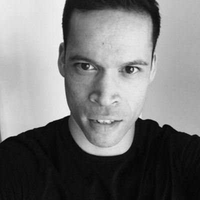 Man in zwart overhemd, close-up, kijkt recht in de camera met neutrale uitdrukking. Zwart-wit.