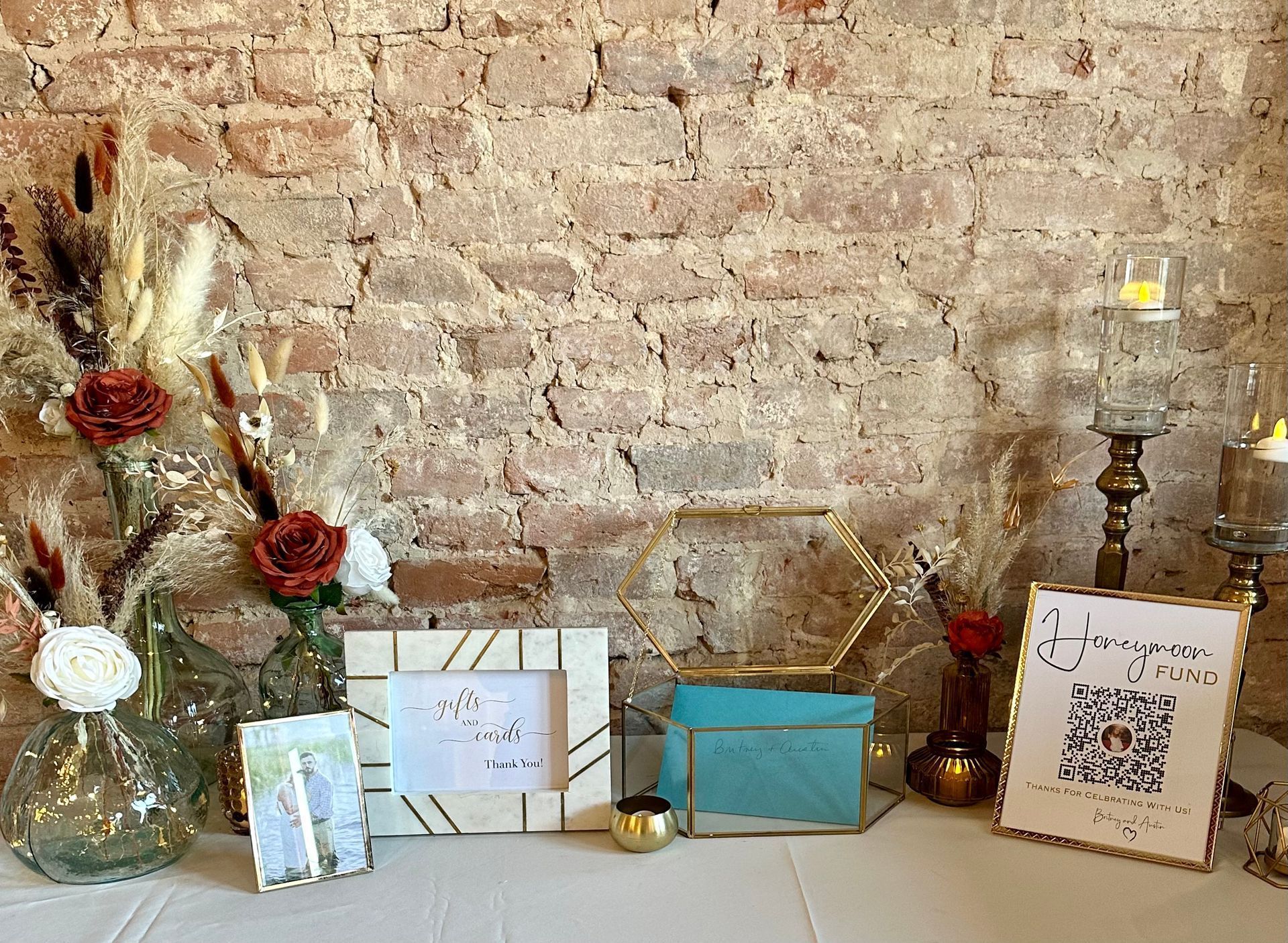 A table with vases , candles , frames , and cards on it in front of a brick wall.