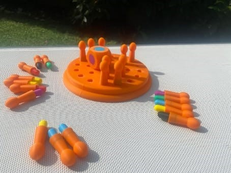 Juego de mesa naranja con clavijas y piezas de colores sobre una superficie blanca.