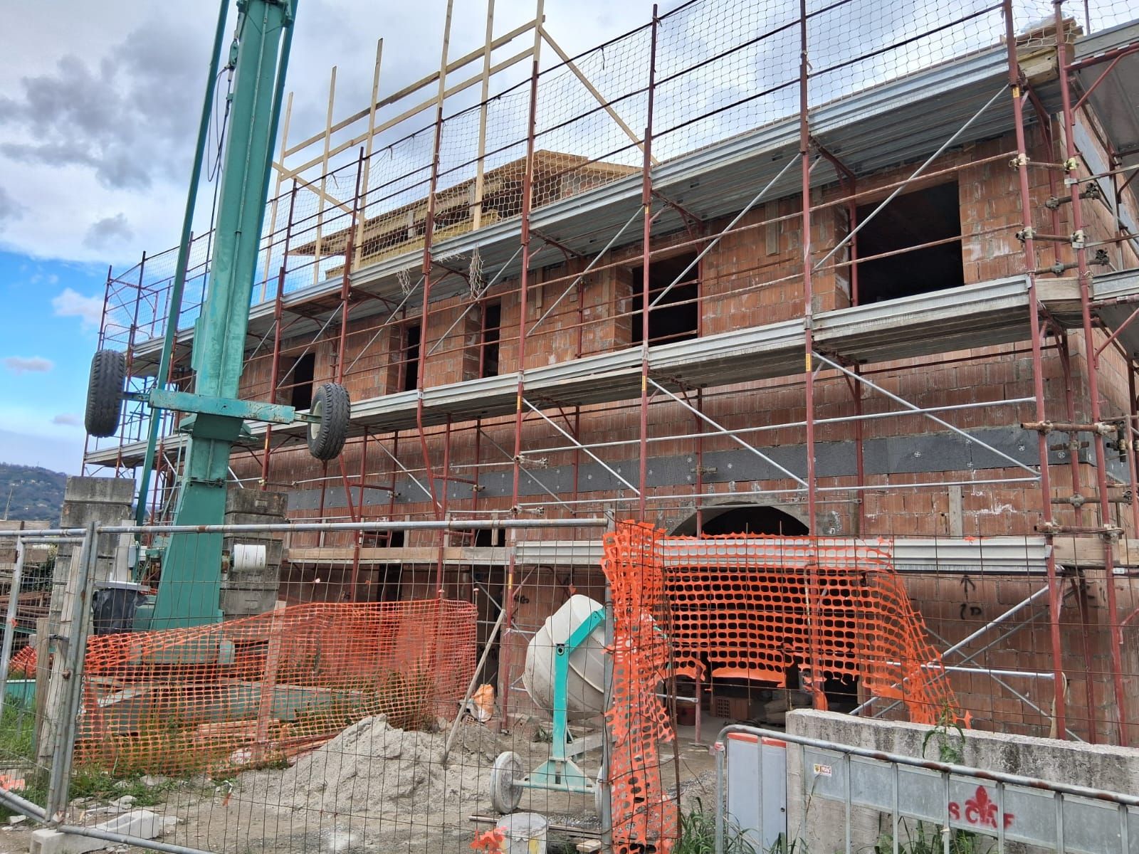 Casa in mattoni in costruzione con impalcature, rete di sicurezza arancione e un operaio in primo piano.