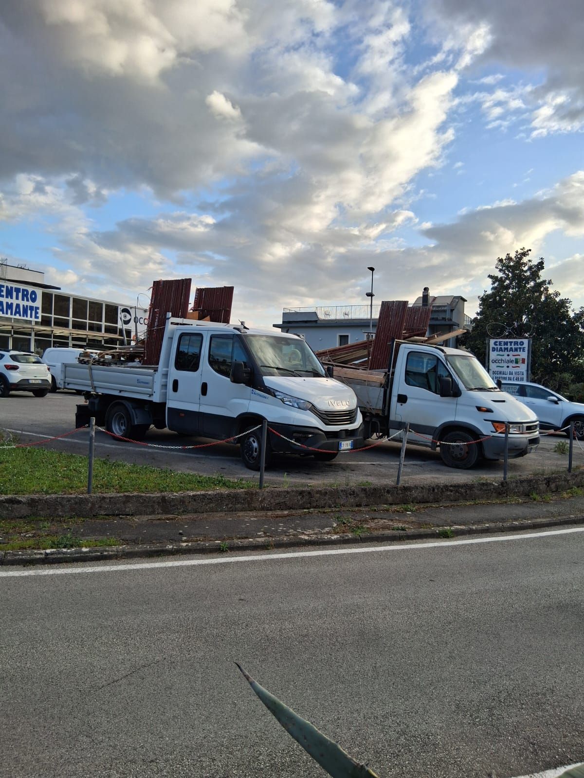 Camion di servizio bianchi parcheggiati a bordo strada sotto un cielo nuvoloso, vicino a un parcheggio e ad alcuni edifici.