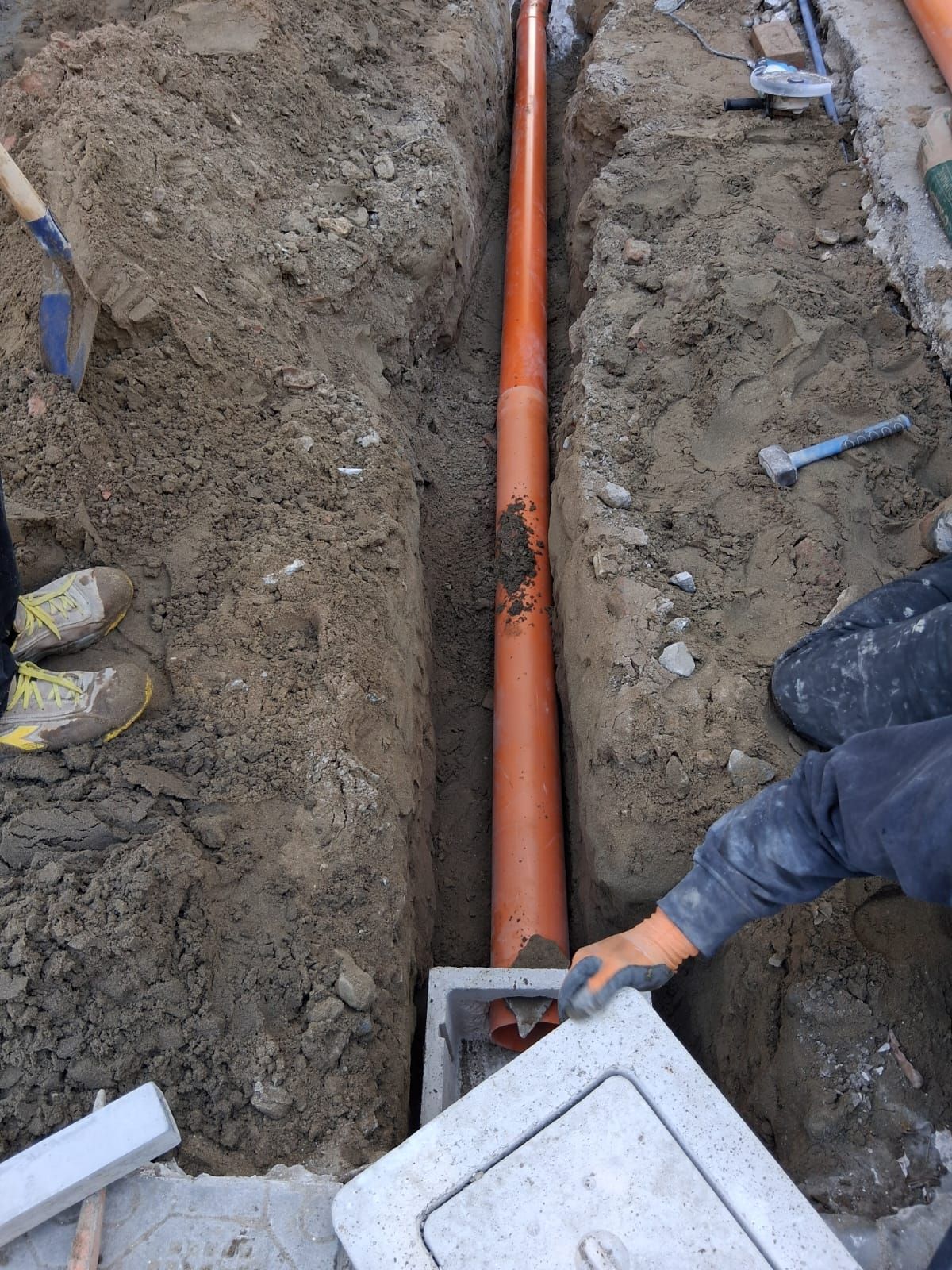 Operai intenti a installare un tubo arancione in una stretta trincea, con il coperchio di una scatola di derivazione in cemento nelle vicinanze.