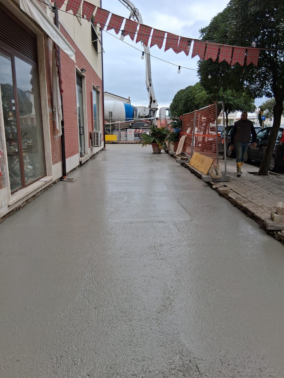 Marciapiede cittadino bagnato accanto agli edifici, con bandierine rosse sopra e barriere di cantiere più avanti.