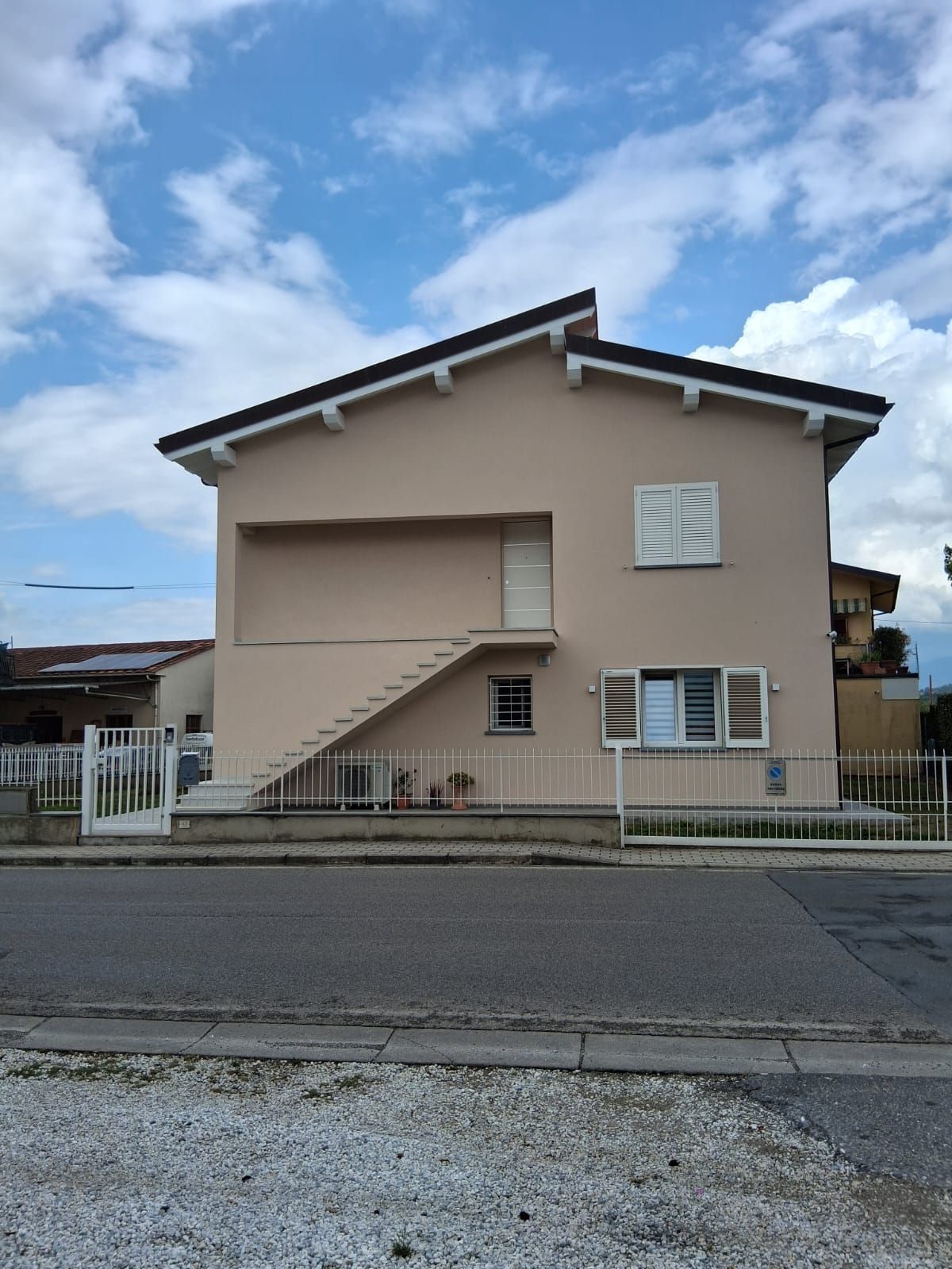 Casa beige a due piani con tetto spiovente, recinzione anteriore e strada antistante sotto un cielo nuvoloso.