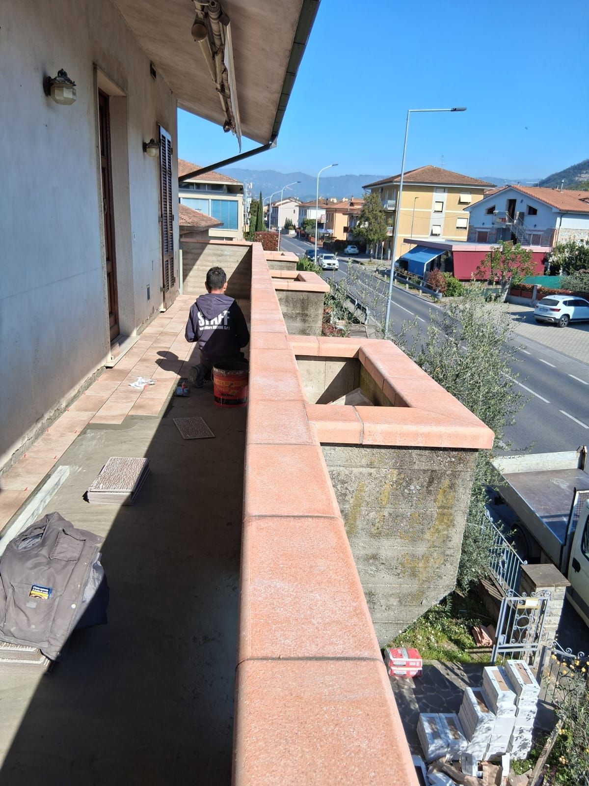Una persona seduta sul balcone stretto di un appartamento, affacciato su una strada, in una giornata di sole.