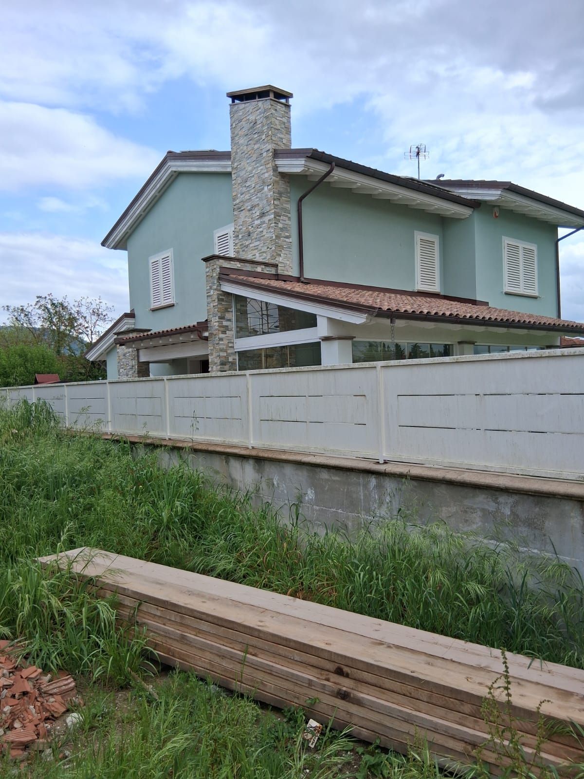 Casa a due piani color verde menta dietro una recinzione bianca, con tetto di tegole rosse e marciapiede erboso antistante.