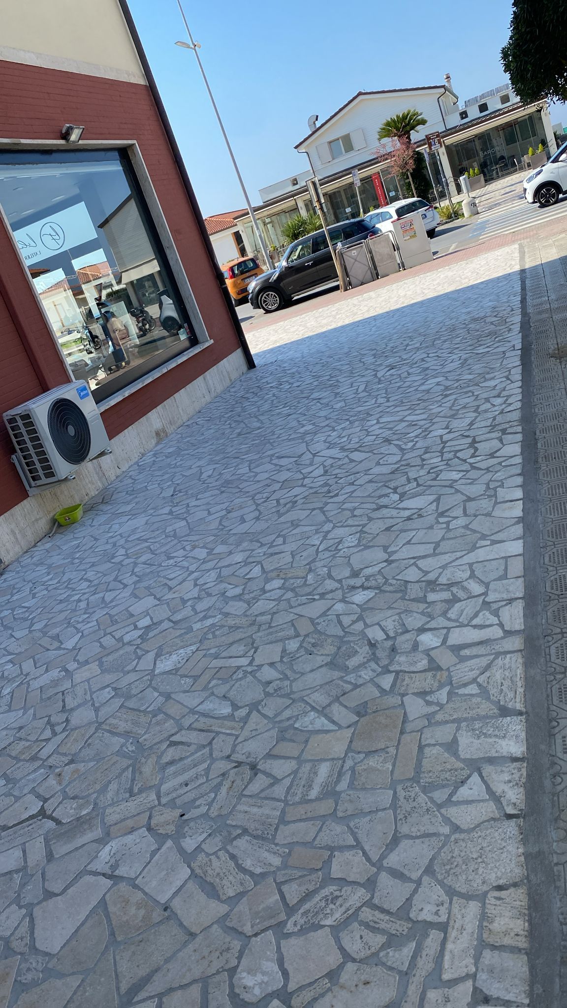 Un passaggio lastricato in pietra, adiacente a un edificio rosso, conduce a una strada soleggiata con auto parcheggiate e negozi.