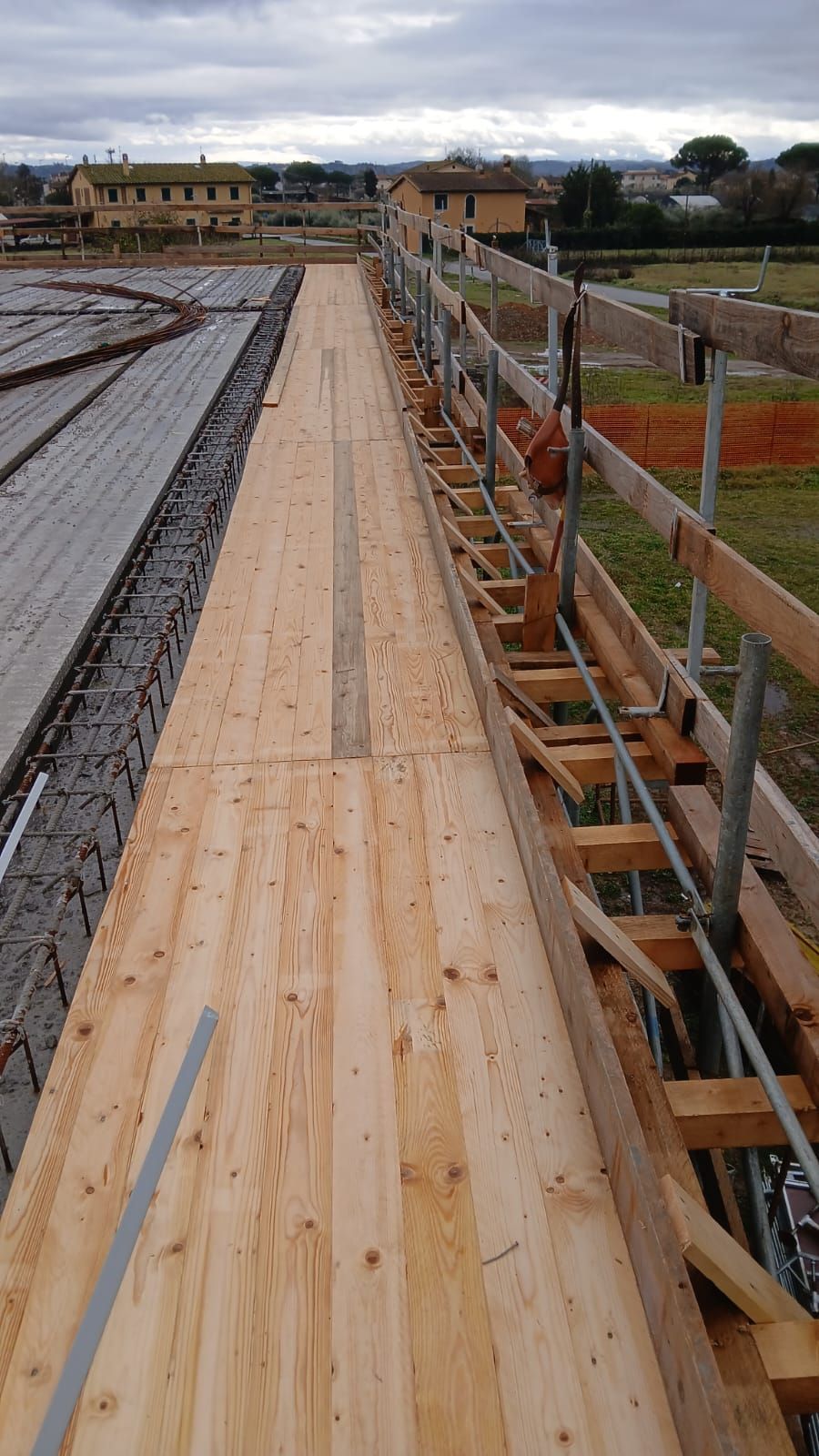 Passerella in legno in un cantiere edile, accanto alla struttura del tetto e a una rete di sicurezza arancione.