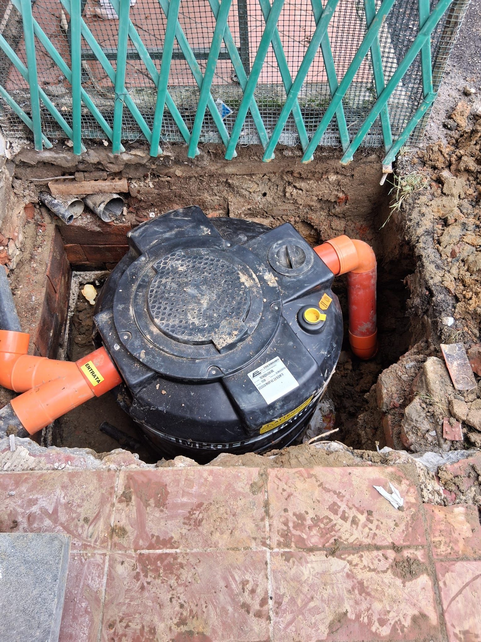 Camera di servizio sotterranea nera con tubature arancioni accanto a un vialetto di mattoni e una recinzione verde.