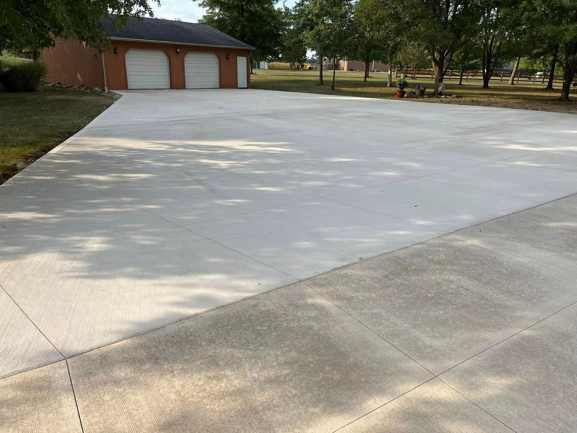 Wide, light gray concrete driveway leading to a garage with two white doors, with grass and trees in the background.
