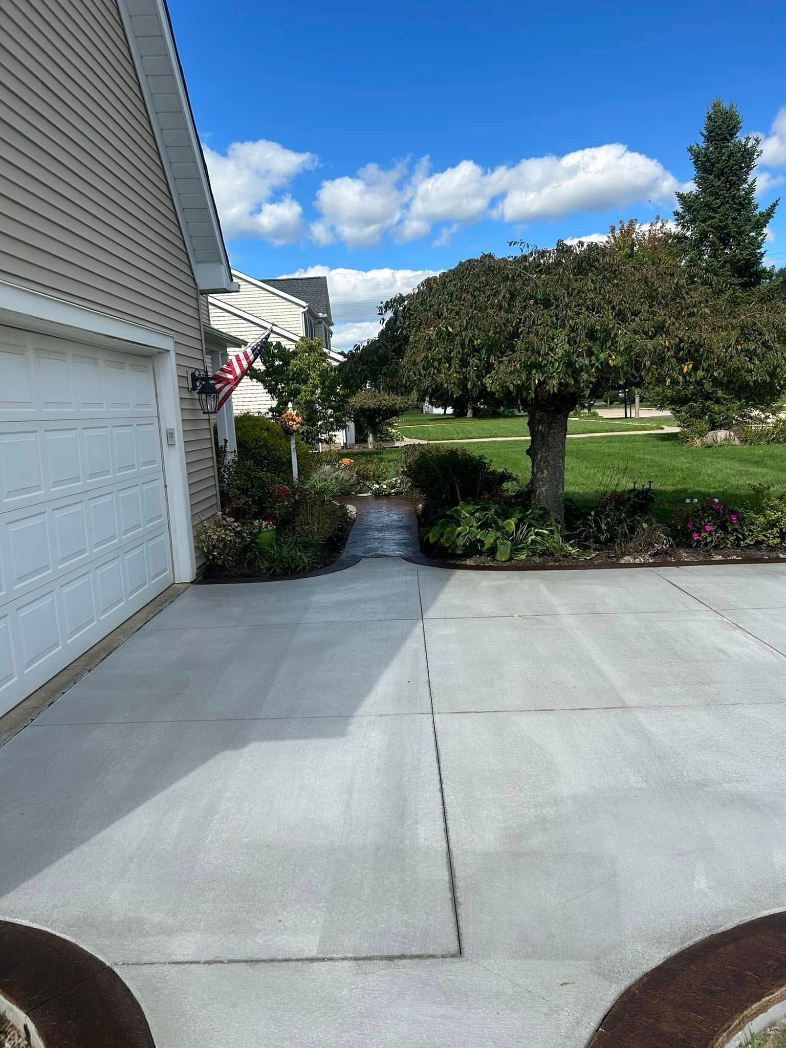 Driveway leads to garden with trees, bushes, and a blue sky with clouds. A garage is on the left.