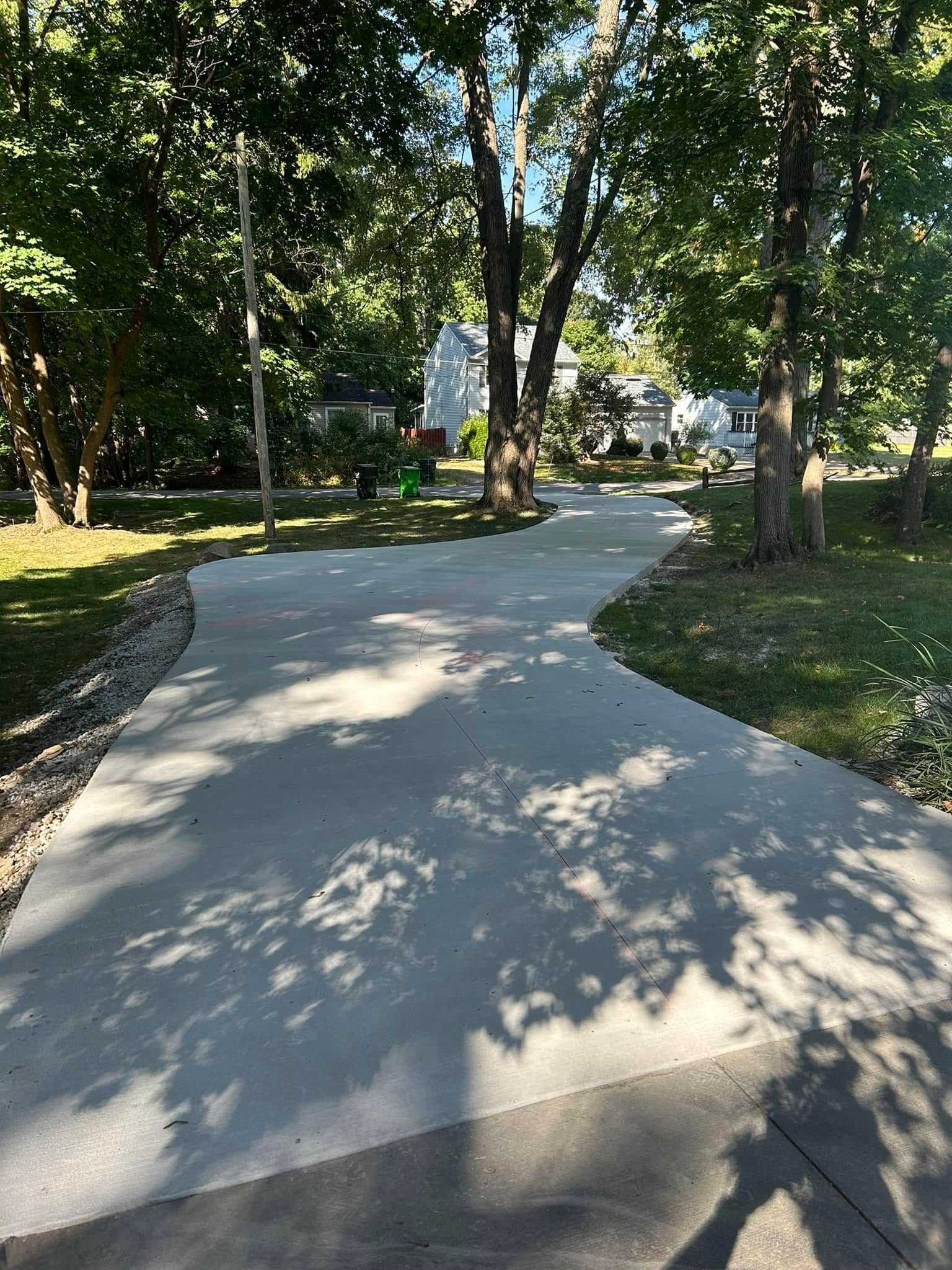 Concrete driveway curves through a tree-lined yard towards a house in the distance; sunny day, shadows.