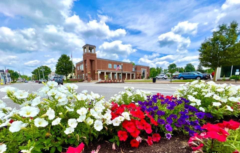 Flowers line a street, leading to a brick building with a clock tower under a cloudy blue sky.